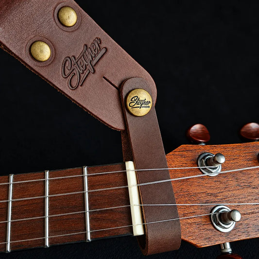 Hochwertiger Gurtverbinder für Ukulele und Mandoline aus dunkelbraunem Echtleder mit Steyner-Logo auf der Alt-Messing-Schraube, hinter dem Sattel einer Ukulele befestigt