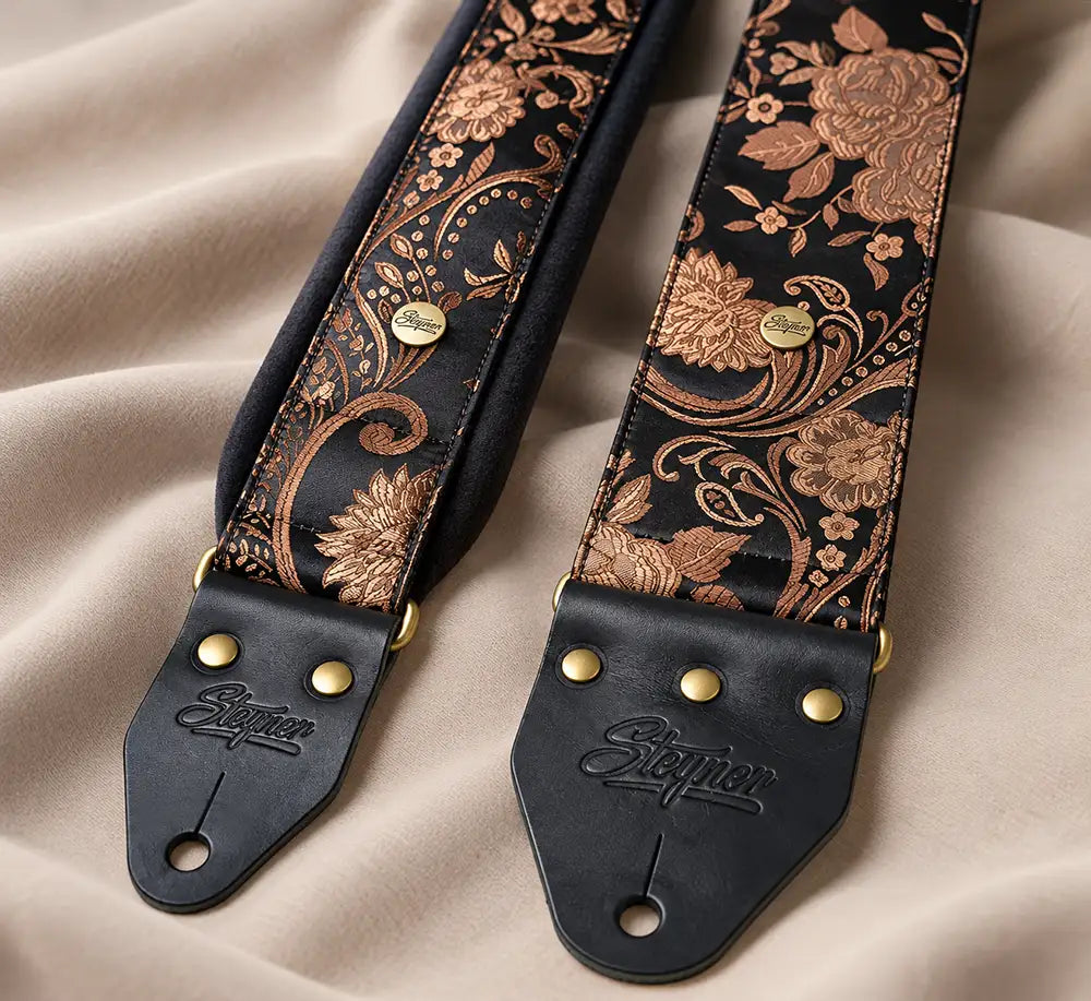 Vergleich zweier handgefertigter Steyner-Gitarrengurte: links ein gepolsterter Gurt, rechts ein extra breiter 8-cm-Gurt, beide mit schwarz-goldenem Blumenmuster und Lederenden.
